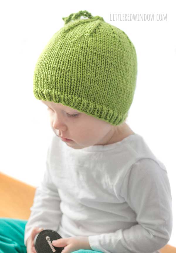 baby in white shirt wearing a bright green knit hat with curly tendril on top that looks like a sweet pea looking down off to the left