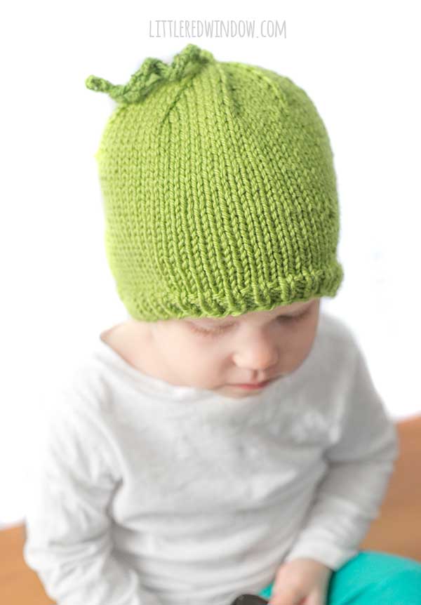 baby in white shirt wearing a bright green knit hat with curly tendril on top that looks like a sweet pea looking slightly to the right