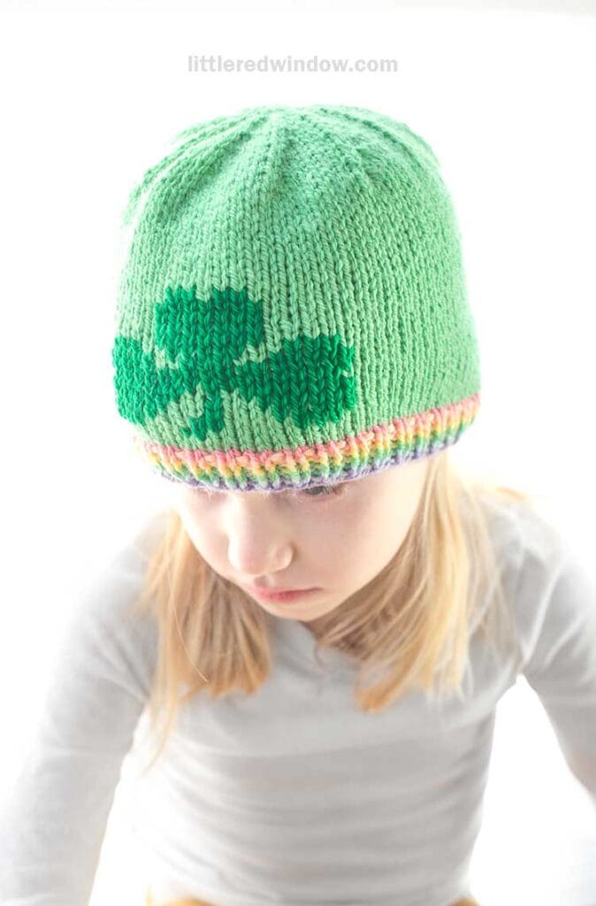 A child is sporting a green knit hat adorned with a shamrock design, showcasing a colorful rainbow brim. The little one gazes downward, dressed in a light gray striped shirt. The vibrant, blurred background adds to this charming scene inspired by the shamrock hat knitting pattern.
