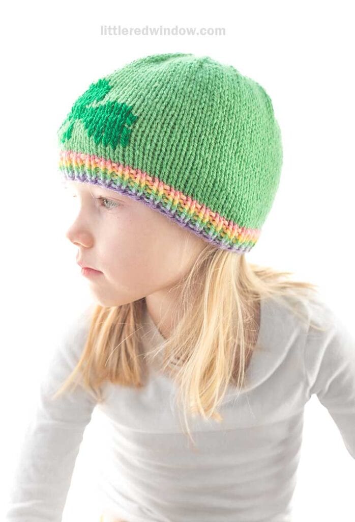 A young child with blonde hair wears a charming shamrock hat featuring a leaf design and a multicolored rainbow brim, looking to the side against a plain white background. She is dressed in a light gray long-sleeve shirt, embracing both style and comfort.