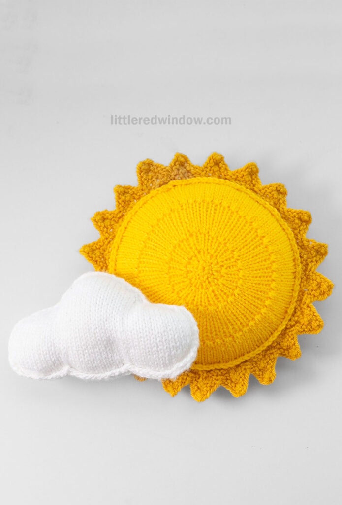 bright yellow knit stuffed sun shape with a knit white cloud shape in front of it on a light fray background