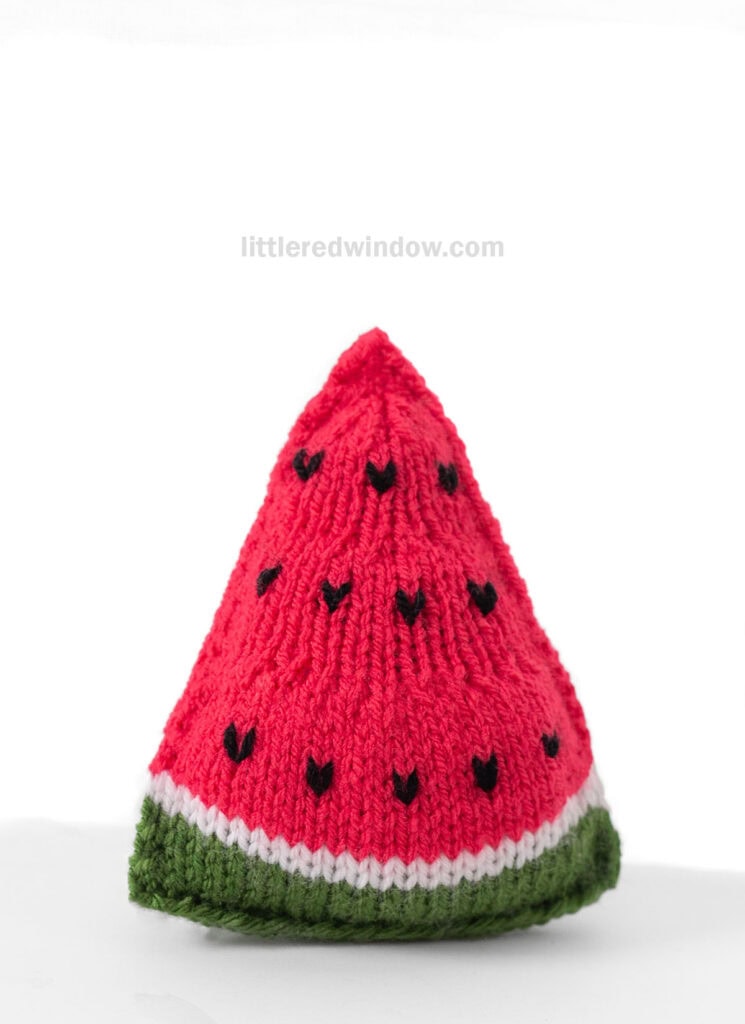 triangular knit watermelon slice with green rind and bright pink watermelon with black seeds from the front on a white background