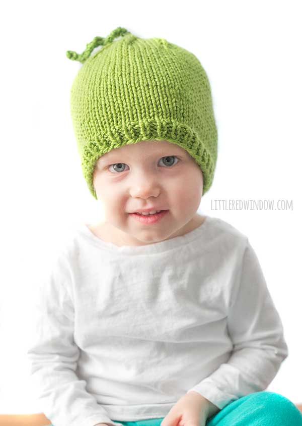 baby in white shirt looking at the camera while wearing a bright green knit hat that looks like a pea in front of a white background