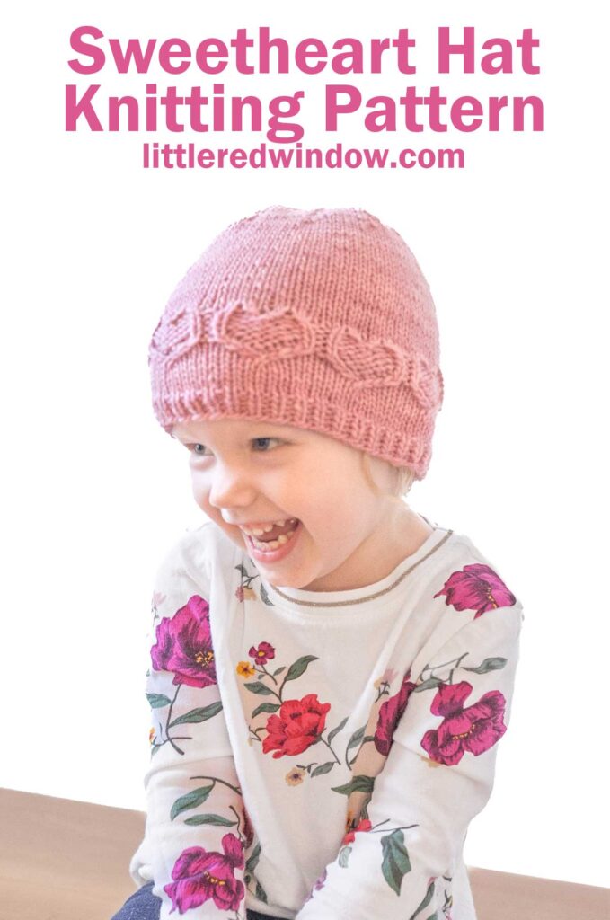 A smiling child dons a pink knitted