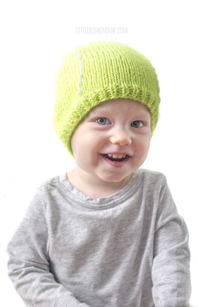 smiling baby in gray shirt wearing bright green tennis ball hat