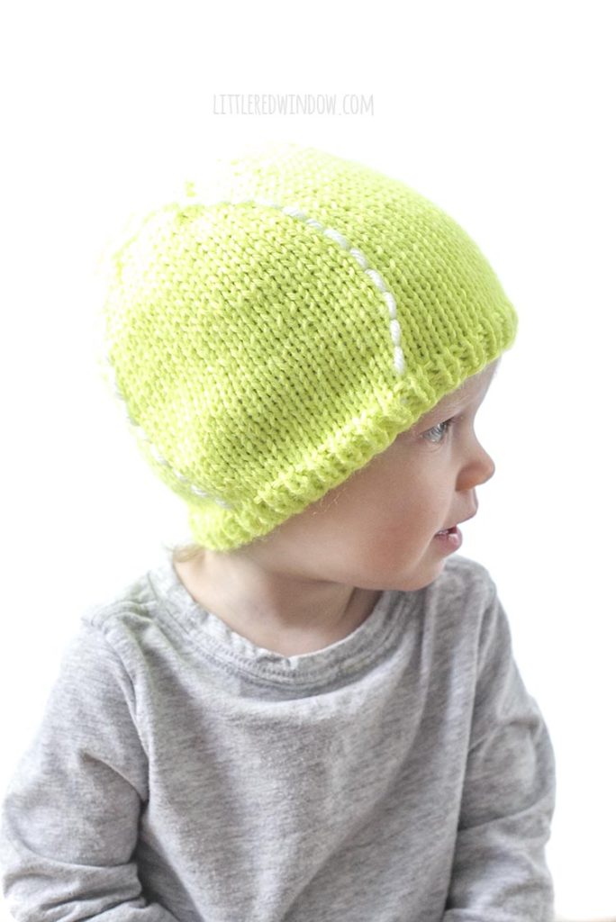 cute baby in gray shirt looking off to the right and wearing a bright green tennis ball hat