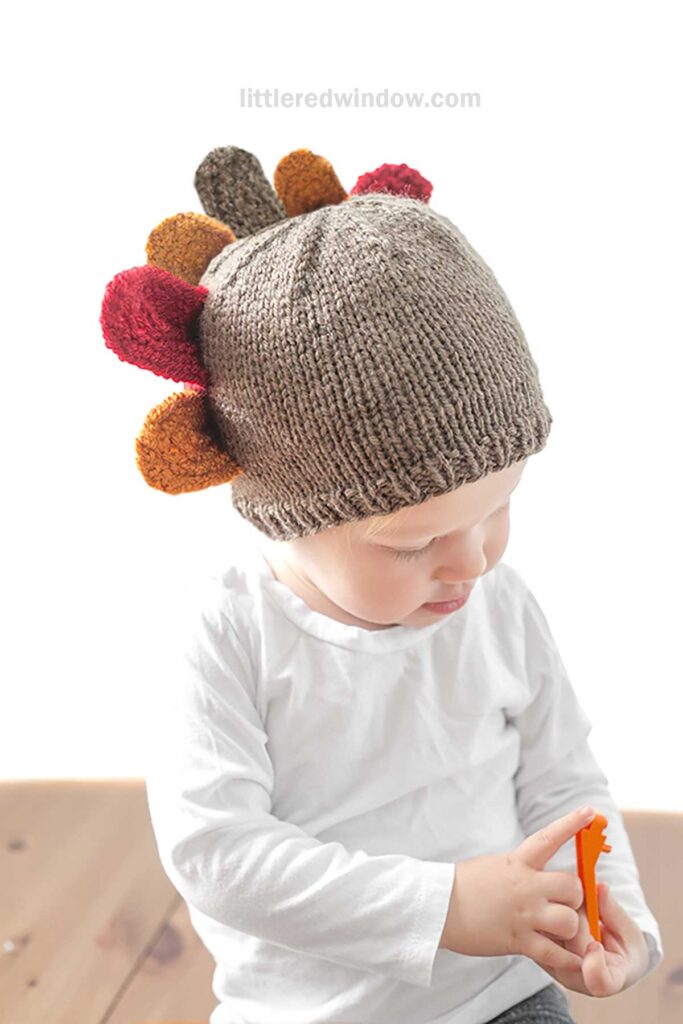 A young child wearing a turkey hat, designed with colorful tail feathers protruding from the top, is dressed in a white long-sleeve shirt. The child looks down while holding an orange object. The background is white and minimalistic.