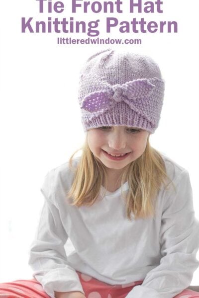 Smiling girl in white shirt and pink pants wearing a light purple knit hat with two side flaps that come together and tie in a bow in the front of the hat looking down at her lap