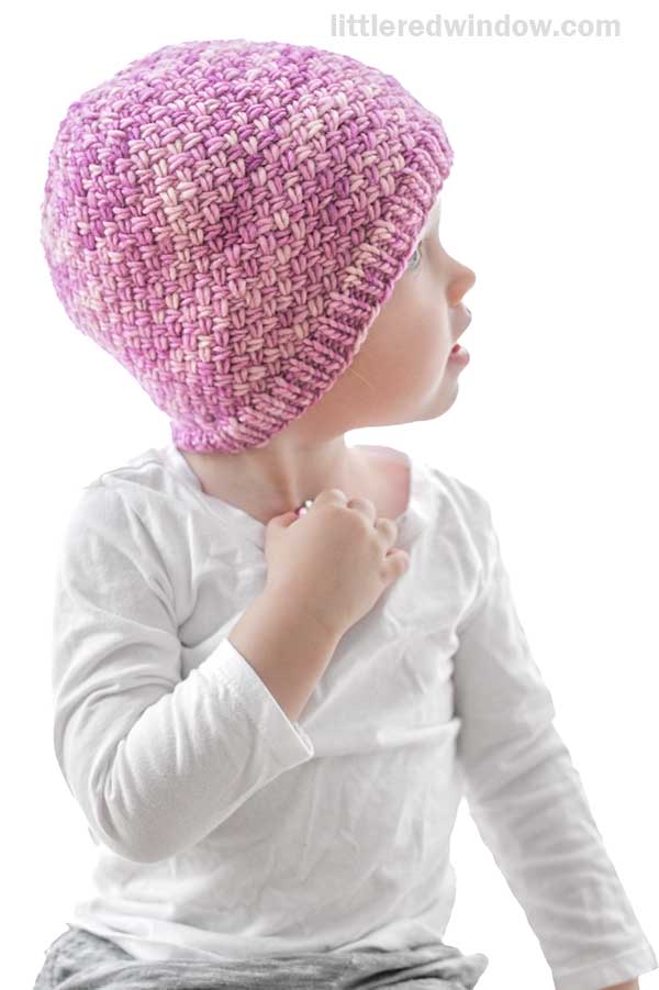A young child wearing a light pink and purple Tiny Basketweave Hat and a white long-sleeve shirt looks to the right against a white background.