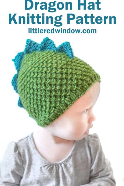 A young child wearing a green, textured dragon hat with blue triangular spikes along the top mimicking a dragon's back. The text above the child reads "Dragon Hat Knitting Pattern" and "littleredwindow.com." The child is dressed in a gray shirt.