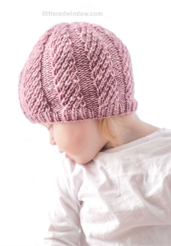 A young child wearing a light pink twisted rib hat looks down. They have on a white long-sleeve shirt. The background is plain and bright, focusing attention on the child.
