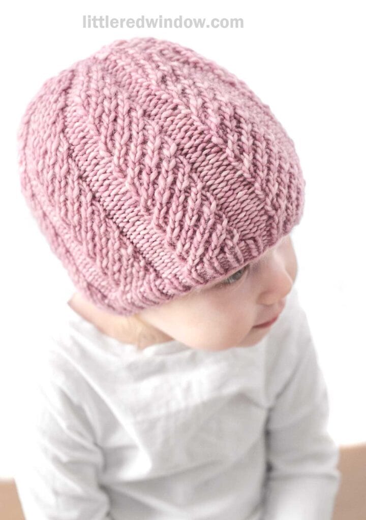 A baby wearing a pink, Twisted Rib Hat looks to the side. The baby is dressed in a white shirt and is positioned against a white background. The hat features a textured pattern. Text at the top reads