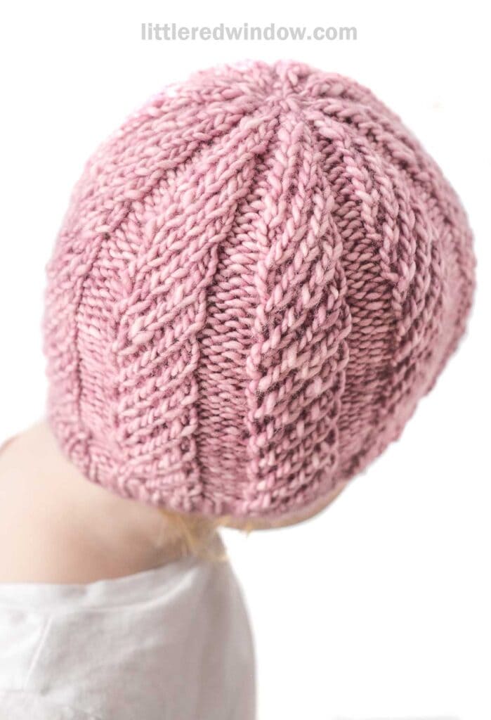 A close-up image of the back of a person's head wearing a pink twisted rib hat with a braided pattern. The person is wearing a white top, and the background is plain white.