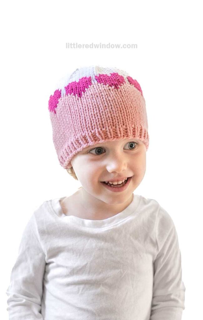 A smiling child wearing a pink and white colorblock knit hat adorned with a row of pink hearts near the top. The child is dressed in a white long-sleeve shirt, set against a plain white background, showcasing an adorable knitting pattern.