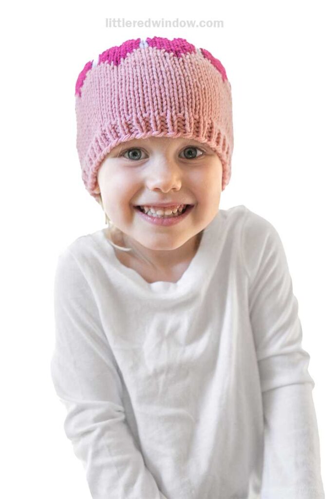 A smiling child in a white shirt wears a charming colorblock knit heart hat embellished with hearts. The background is plain white.