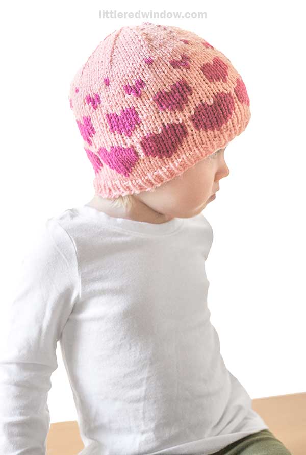 A child sits sideways, wearing a white shirt and a pink knit hat adorned with a charming red hearts pattern. The background is neutral and plain.