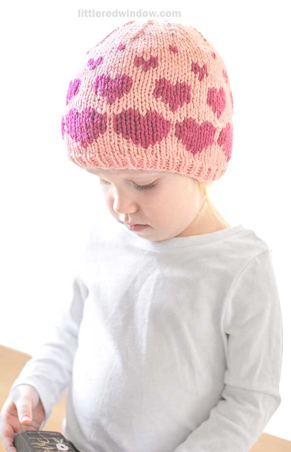 A child wearing a knitted pink hat adorned with purple hearts stands looking down slightly, their white shirt bright against the vibrant background.