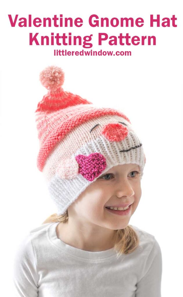 smiling girl in white shirt in front of a white background wearing a knit hat that looks like a smiling white bearded Valentine's Day gnome holding a hot pink heart in his hand and wearing a stocking cap