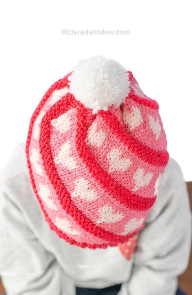 Person wearing a pink and red knitted swirl hat with white heart patterns. The hat is topped with a fluffy white pom-pom. The individual is facing downward against a white background.