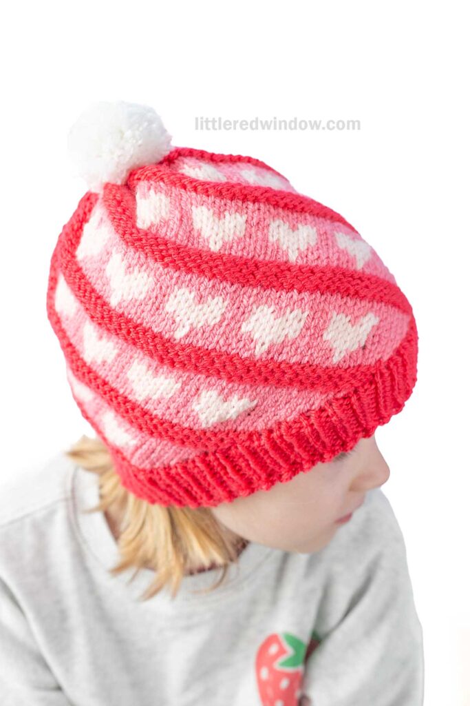 Child wearing a red and pink knit swirl hat with white hearts and a white pom-pom on top. The child is facing sideways, and the sweater has a small strawberry design. The background is plain white.
