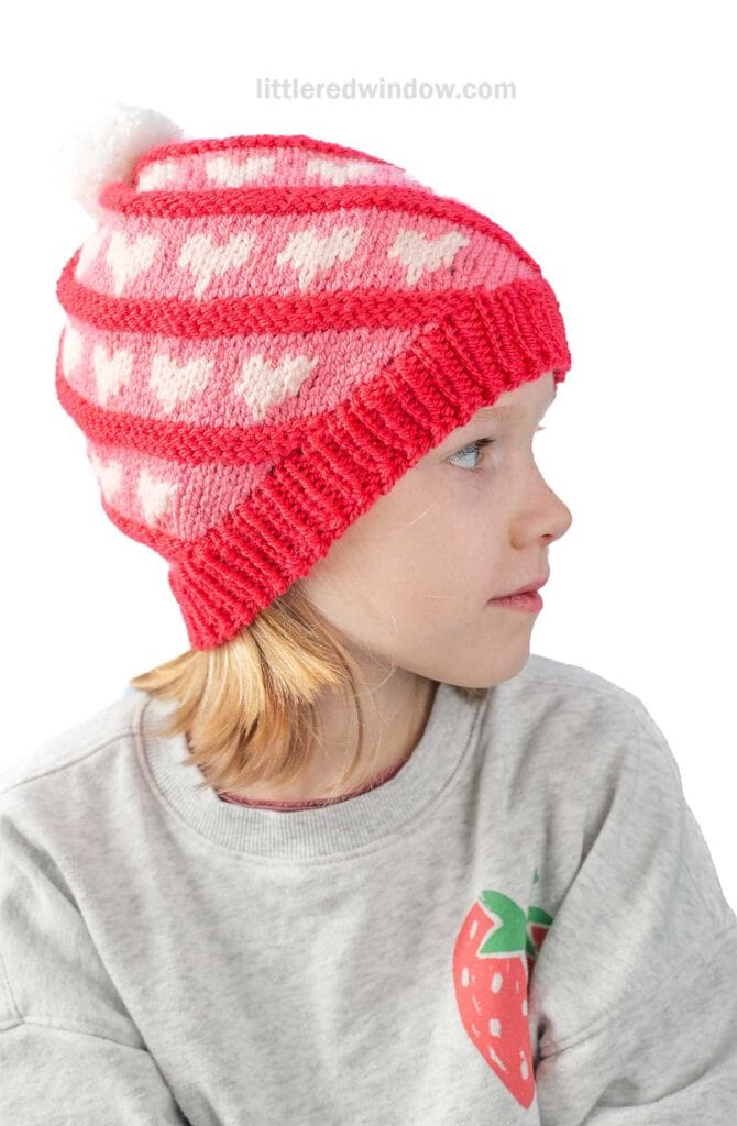 A child wearing a gray sweatshirt with a strawberry design looks to the side. They have a red and pink knit hat with a white heart pattern and pom-pom on top.
