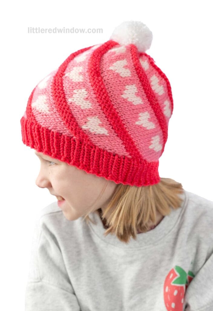 A child wearing a pink and red knitted swirl hat with white hearts and a pom-pom on top. The child is looking to the side, and is dressed in a gray sweater with a strawberry design on the shoulder.