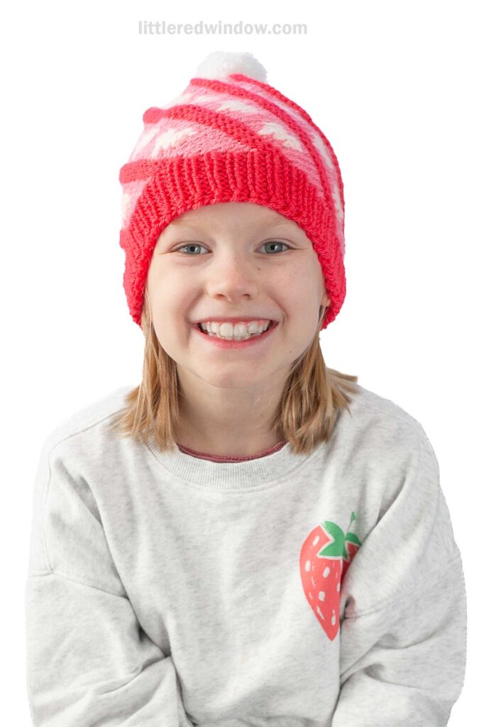 A young girl smiles brightly, wearing a red and white knitted swirl hat with light pink hearts on it and a sweatshirt with a strawberry illustration. The background is plain white.