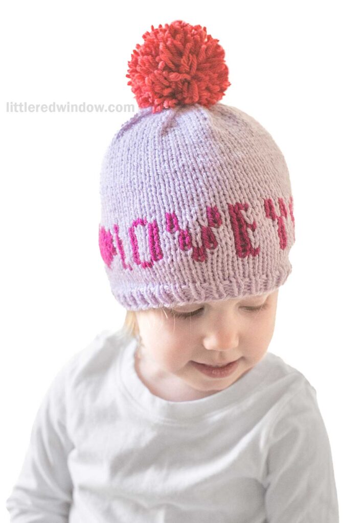 A child wears a Love Notes hat with a red pom-pom on top. The hat is light purple, featuring the words