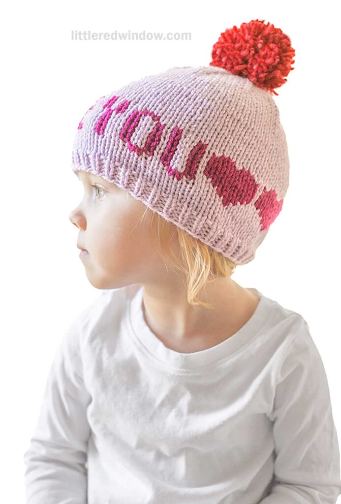 A child in a white shirt gazes sideways, donning a pink knit hat from the Love Notes Hats collection, featuring a red pompom and the words
