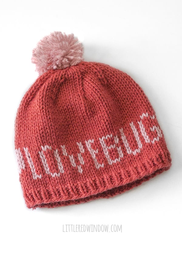 Medium red knit hat with light pink pom pom and light pink words that say LOVEBUG on a white background
