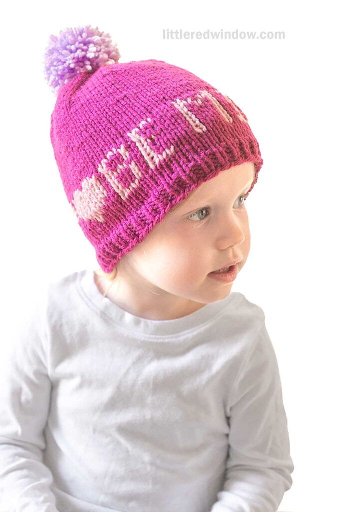 A child wears a pink knit Love Notes Hat, complete with a purple pom-pom and the words