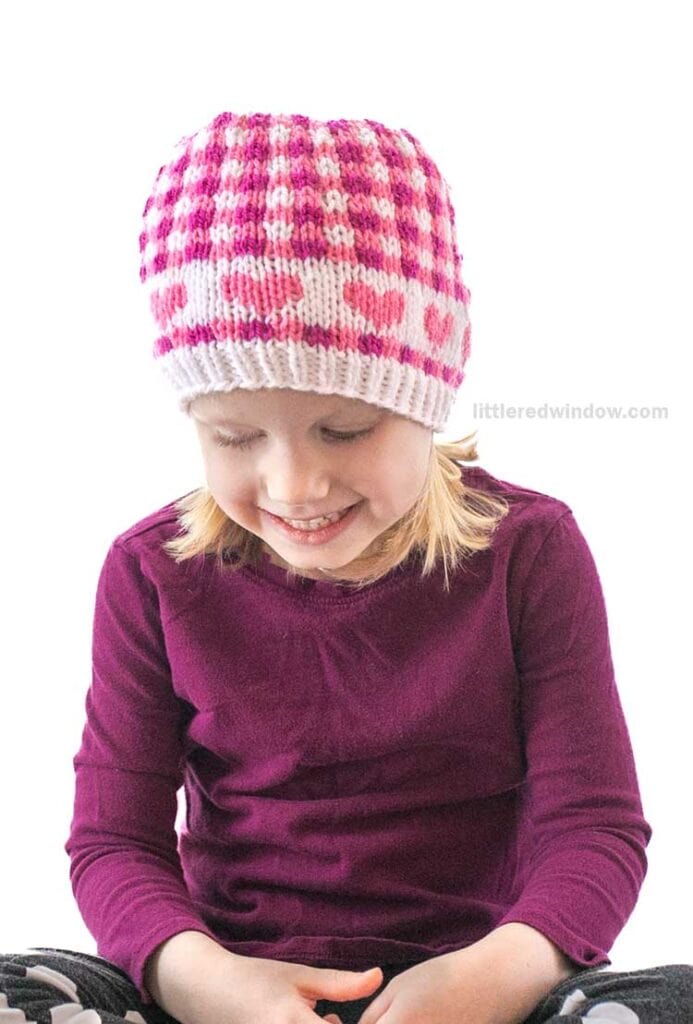 A child wearing a pink and white knitted hat smiles while looking down. Dressed in a long-sleeve burgundy shirt with subtle Valentine plaids, they sit with hands in lap against a plain white background.