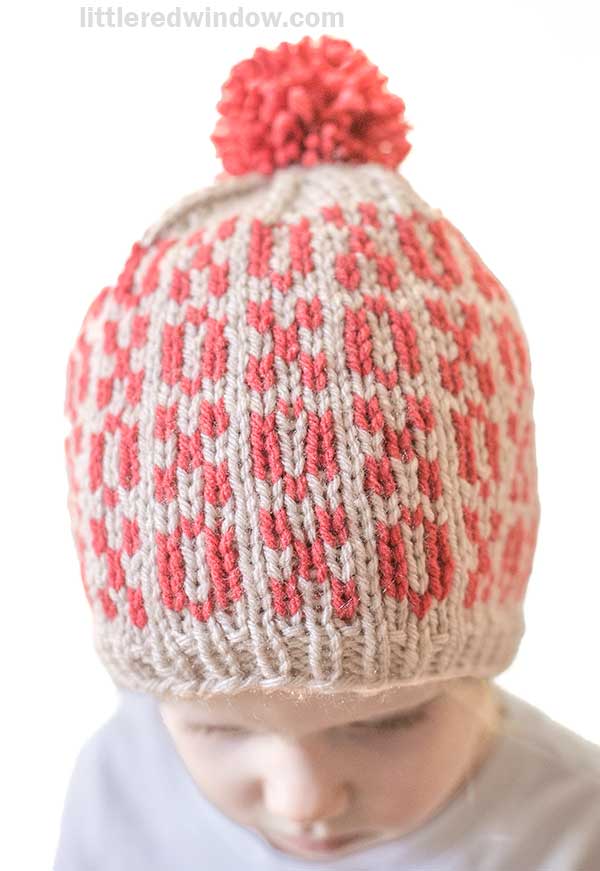 A child grins under a knitted Tic Tac Toe Hat, adorned with a charming red and beige xoxo pattern. The vibrant red pom-pom on top adds a playful touch against the plain white background.