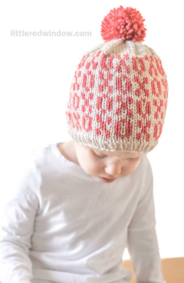 A child wearing a Tic Tac Toe Hat with a red and cream checkerboard xoxo pattern and a playful red pom-pom on top, dressed in a white shirt, gazes downward thoughtfully.