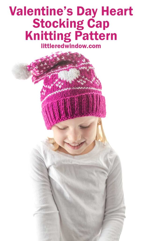 A child wearing a pink knitted Heart Stocking Cap with hearts and a pom-pom, smiles while looking down. Text above reads,