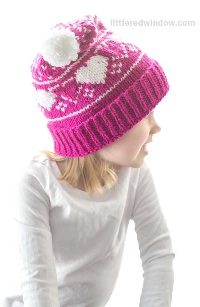 A child in a white shirt is smiling and looking to the side, wearing a bright pink heart stocking cap adorned with cute white heart patterns and a playful pom-pom on top.