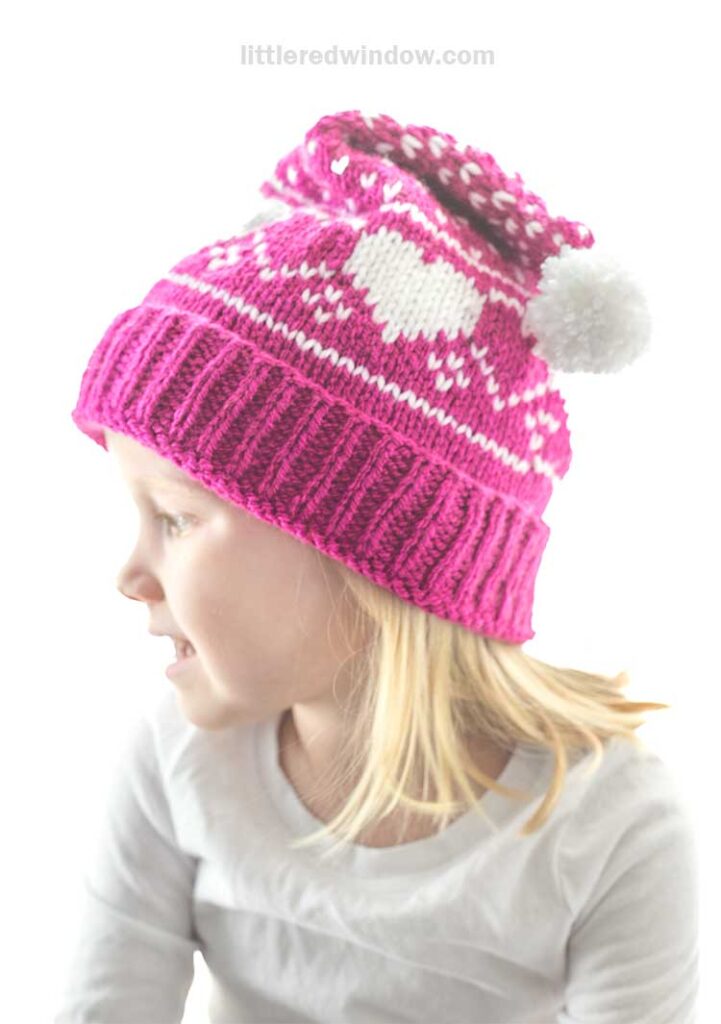 A child sporting a heart stocking cap, bright pink with white heart patterns and topped with a fluffy pom-pom, gazes to the side in their white shirt. The soft gradient background enhances the light and airy ambiance of the scene.