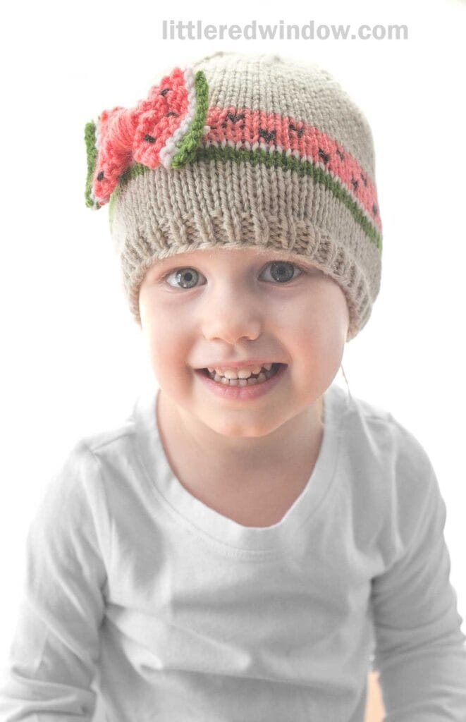 A smiling young child is wearing a beige bow hat adorned with a pink and green decorative pattern, showcasing intricate knitting. The child is dressed in a white shirt and is looking directly at the camera. The text
