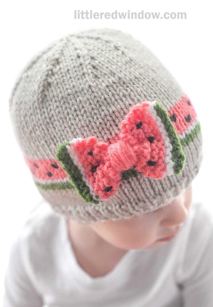 A baby is wearing a hand-knitted gray bow hat adorned with a colorful design of a pink and green bow resembling a watermelon slice. The website 