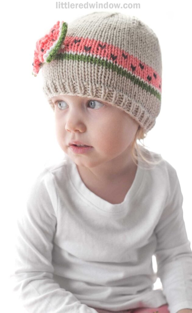 A young child wearing a white long-sleeve shirt and a knitted bow hat with a pink, green, and red floral pattern sits and looks to the side. The background is plain and light-colored.