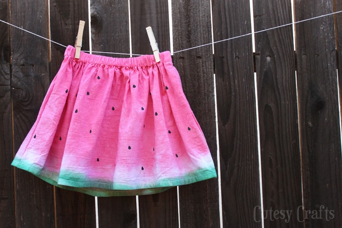 Childs skits dyed pink with green hem to look like a watermelon hanging on a clothesline in front of a dark wood fence