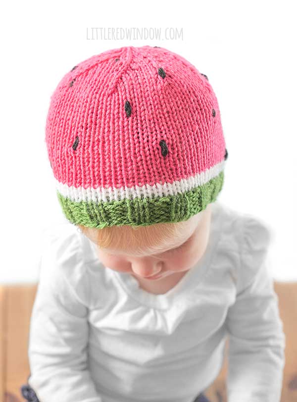 baby wearing a fun knit hat that looks like a watermelon slice complete with seeds looking down to the left