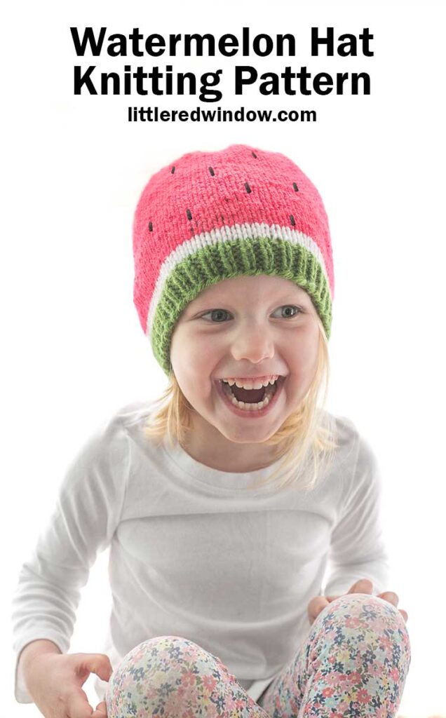 laughing child in white shirt wearing a knit watermelon hat in green white and pink with black knit seeds in front of a white background