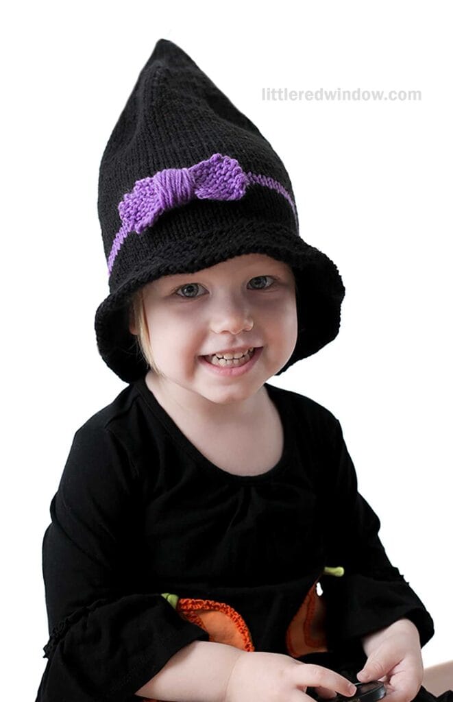 A young child smiles while wearing a black outfit and a tall, pointed witch hat adorned with a purple bow. They are seated and looking directly at the camera. The background is a plain white.