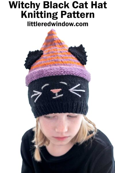 A person is wearing a knitted hat that looks like a black cat with a striped purple and orange top section. The hat features black cat ears, white whiskers, a pink nose, and white eyes. The text above the image reads "Witchy Black Cat Hat Knitting Pattern" from littleredwindow.com.