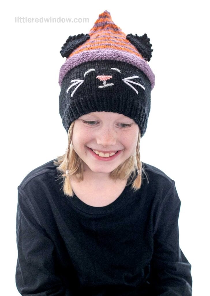 A smiling child with light hair wears a knitted hat designed to resemble a cat. The hat is primarily black with an orange and purple striped section towards the top, adorned with cat ears, white whiskers, and a pink nose. The child is dressed in a black top.