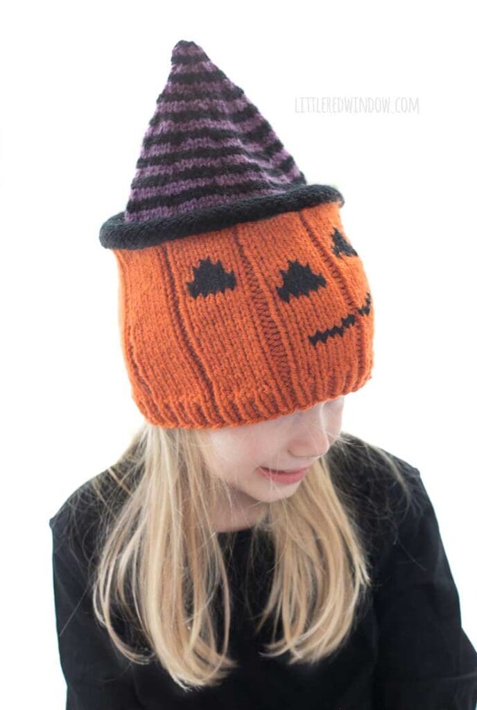 Smiling blond girl wearing a knit hat that looks like a jack o lantern wearing a purple and black striped witch hat while she is is looking down and to the right