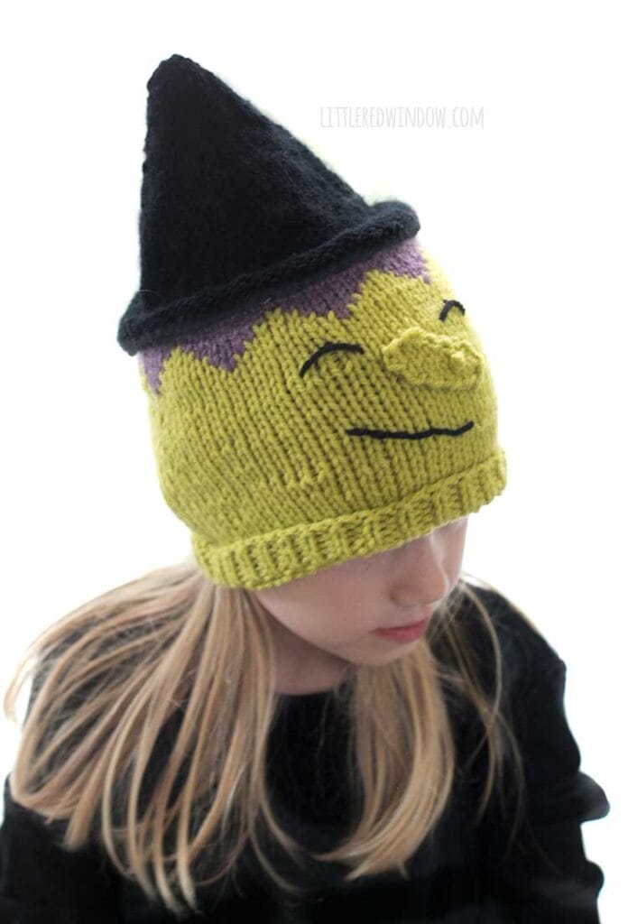 Little blond girl in a black shirt wearing a hat that looks like a witch including green witchy skin warty nose purple hair and black pointy witch hat while the girl is looking down and to the right