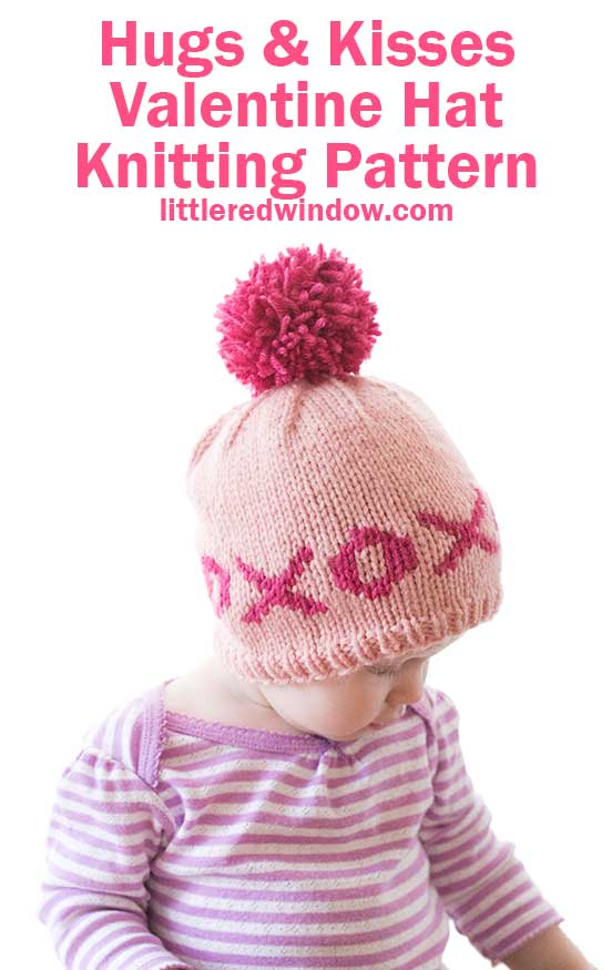 A baby in a striped shirt wears a pink knitted Valentine hat adorned with a red pom-pom and