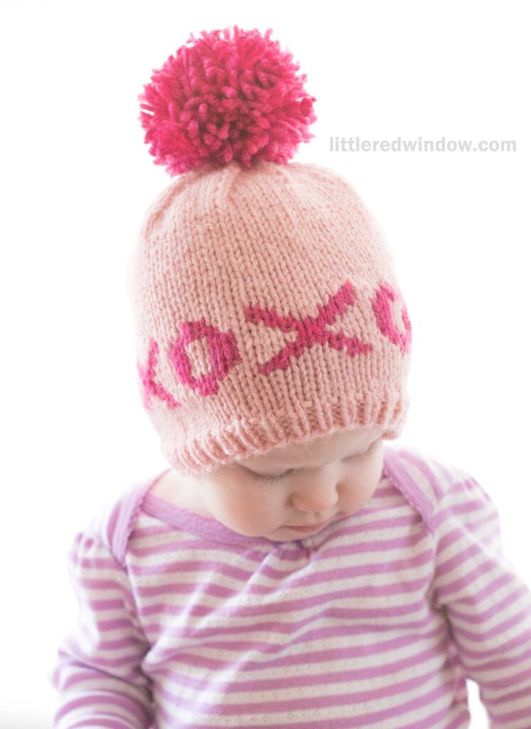 A baby in a pink and purple striped shirt looks down, wearing an adorable valentine hat—a pink knitted cap with a red pom-pom and charming